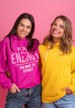 two pretty women friends embracing having fun together, blond and brunette, wearing hoodie posing on pink background isolated